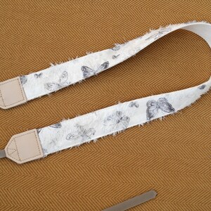 May include: A white camera strap with a butterfly pattern in shades of gray. The strap has a frayed edge and beige leather end pieces with gray connectors. The background is a textured, golden-brown fabric.