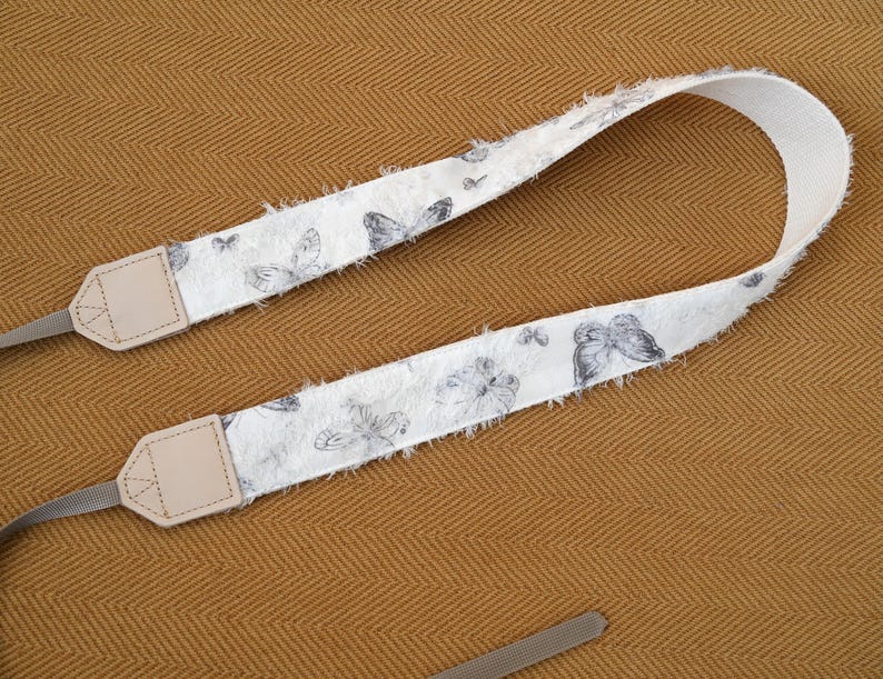 May include: A white camera strap with a butterfly pattern in shades of gray. The strap has a frayed edge and beige leather end pieces with gray connectors. The background is a textured, golden-brown fabric.