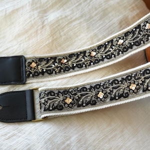 Könnte beinhalten: Ein Gitarrengurt mit cremefarbenem Hintergrund und schwarzer Blumenstickerei. Der Gurt hat schwarze Lederenden und goldfarbene Hardware. Das florale Design umfasst goldene Pailletten und Akzente.