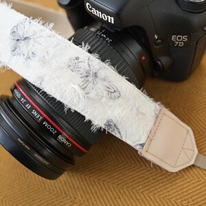 May include: A Canon EOS 7D camera with a white camera strap featuring a butterfly pattern. The strap has a frayed edge and a tan leather end piece. The lens is a Canon Zoom Lens EF 24-105mm.
