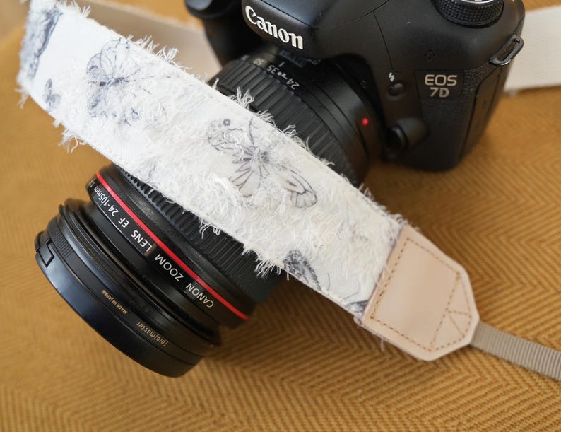 May include: A Canon EOS 7D camera with a white camera strap featuring a butterfly pattern. The strap has a frayed edge and a tan leather end piece. The lens is a Canon Zoom Lens EF 24-105mm.