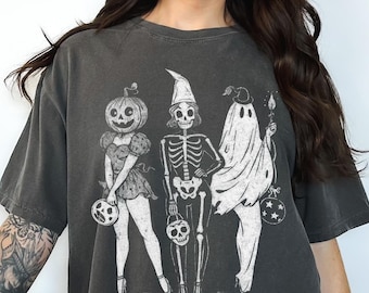 Puede incluir: Camiseta gris oscuro de gran tamaño con un diseño de Halloween. El diseño muestra un personaje con cabeza de calabaza, un esqueleto y un fantasma, cada uno sosteniendo una pequeña calabaza o linterna. La camiseta tiene un aspecto vintage.