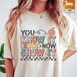 May include: A white t-shirt with the text "YOU KNOW IT NOW SHOW IT" in colorful letters with a peach, pencil, and stars. The text is surrounded by a black and white checkered border.