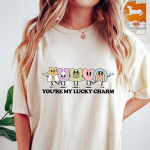 May include: A cream colored t-shirt with a graphic of five cartoon lucky charms. The charms are a star, horseshoe, four-leaf clover, heart, and rainbow. The text "YOU'RE MY LUCKY CHARM" is printed below the charms.