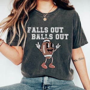 May include: Dark grey t-shirt featuring the text "FALLS OUT BALLS OUT" above a cartoon football character. The football character is giving the middle finger. The t-shirt is worn with blue denim jeans.