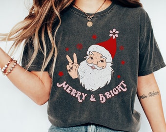May include: Dark grey t-shirt with a Santa Claus graphic, peace sign, and the pink text "Merry & Bright". Santa wears a red hat. The shirt is worn with blue jeans and white trainers.