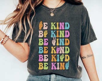 May include: A dark grey t-shirt with the phrase "Be Kind" repeated five times in colourful letters and hand gestures. The shirt is worn by a person with long blonde hair and blue jeans.