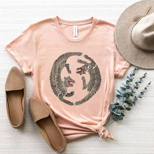 May include: A peach-coloured t-shirt featuring a black and white graphic of two tigers facing each other in a circle. The shirt is made from a soft, comfortable material and has a relaxed fit.