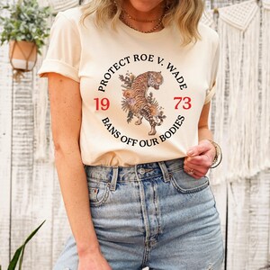 Puede incluir: Una camiseta de color crema con un estampado gráfico de un tigre con detalles florales. El gráfico también incluye el texto "Protect Roe v. Wade 1973 Bans Off Our Bodies".