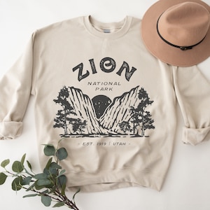 K&ouml;nnte beinhalten: Beiges Sweatshirt mit schwarzem Grafikdesign des Zion National Park Logos. Das Design enth&auml;lt den Text "ZION NATIONAL PARK" &uuml;ber einer Berglandschaft. Ein brauner Hut mit schwarzem Band liegt obenauf.