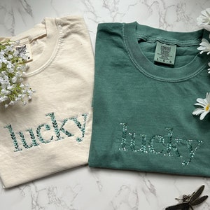 May include: Two t-shirts with the word "lucky" embroidered in green leaves on a cream and a teal shirt.
