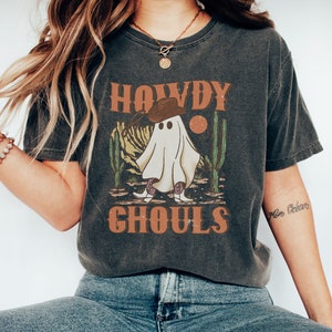 K&ouml;nnte beinhalten: Ein dunkelgraues T-Shirt mit einer Vintage-Grafik eines Geistes, der einen Cowboyhut und Stiefel tr&auml;gt. Der Text "Howdy Ghoulds" ist auf dem Shirt aufgedruckt.