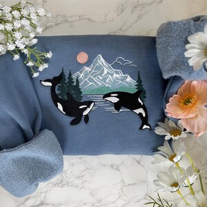 Puede incluir: Una sudadera azul con un dise&ntilde;o bordado de dos orcas nadando frente a una cordillera con un lago y &aacute;rboles. El dise&ntilde;o est&aacute; en hilo blanco, negro y verde.