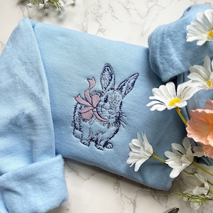May include: A light blue sweatshirt with an embroidered design of a rabbit holding a pink bow. The rabbit is in a light blue color with dark blue outlines.