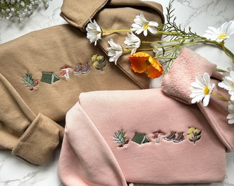 Puede incluir: Dos sudaderas, una color beige y otra rosa, con diseños bordados con temática de camping. La sudadera beige tiene cuello y puños doblados. La sudadera rosa tiene cuello y puños doblados. Los diseños bordados incluyen una tienda de campaña, una seta, botas y follaje.