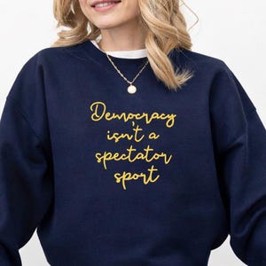 Può includere: Felpa blu navy con la scritta dorata "Democracy isn't a spectator sport". La felpa è abbinata a jeans blu e scarpe da ginnastica nere. È visibile una collana d'oro.