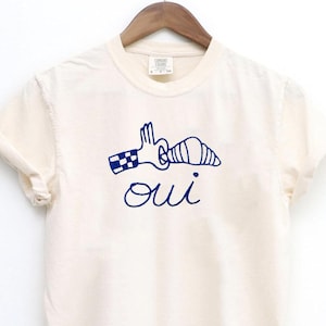 May include: Cream-colored t-shirt with a blue graphic of a hand, croissant, and the word "oui." The t-shirt is hanging on a wooden hanger against a white background. The shirt has rolled-up sleeves.