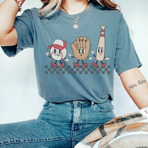 May include: A blue t-shirt featuring a graphic of a baseball, baseball glove, and baseball bat with cartoon faces. The graphic is set against a checkered background. The shirt is being worn by a person with a necklace and a bracelet.