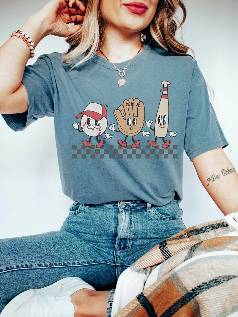 May include: A blue t-shirt featuring a graphic of a baseball, baseball glove, and baseball bat with cartoon faces. The graphic is set against a checkered background. The shirt is being worn by a person with a necklace and a bracelet.