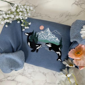 Puede incluir: Una sudadera azul con un dise&ntilde;o bordado de dos orcas nadando frente a una cordillera con un sol rosa en el cielo.