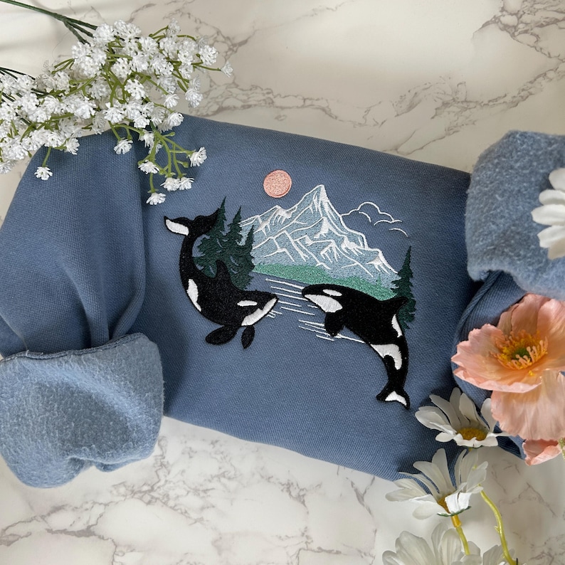 Puede incluir: Una sudadera azul con un dise&ntilde;o bordado de dos orcas nadando frente a una cordillera con un sol rosa en el cielo.