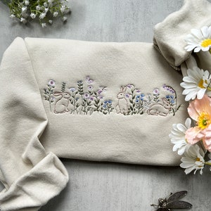 May include: A beige sweatshirt with embroidered bunnies and flowers. The sweatshirt is folded and laid on a gray surface with a dragonfly figurine.
