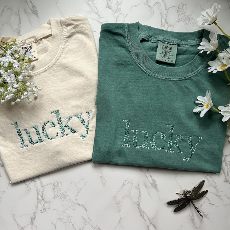 May include: Two t-shirts with the word "lucky" embroidered in green leaves on a cream and a teal shirt.