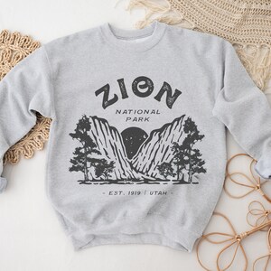 K&ouml;nnte beinhalten: Hellgraues Sweatshirt mit schwarzem Aufdruck des Zion Nationalparks. Der Aufdruck enth&auml;lt den Text "ZION NATIONAL PARK - EST. 1919 - UTAH" mit einem Berg- und Baumdesign. Die &Auml;rmel des Sweatshirts sind hochgekrempelt.