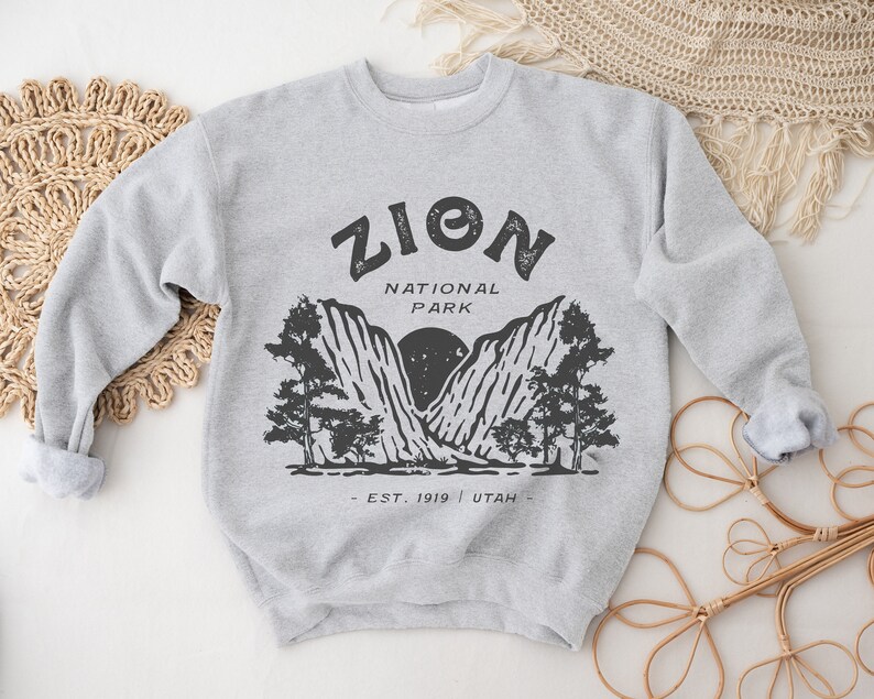 K&ouml;nnte beinhalten: Hellgraues Sweatshirt mit schwarzem Aufdruck des Zion Nationalparks. Der Aufdruck enth&auml;lt den Text "ZION NATIONAL PARK - EST. 1919 - UTAH" mit einem Berg- und Baumdesign. Die &Auml;rmel des Sweatshirts sind hochgekrempelt.