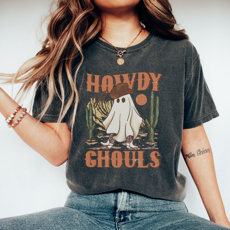 K&ouml;nnte beinhalten: Ein dunkelgraues T-Shirt mit einer Vintage-Grafik eines Geistes, der einen Cowboyhut und Stiefel tr&auml;gt. Der Text "Howdy Ghoulds" ist auf dem Shirt aufgedruckt.