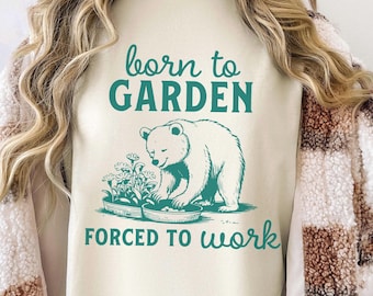 Può includere: Una felpa bianca con il testo "born to GARDEN forced to work" in verde turchese. Un orso polare dei cartoni animati è mostrato mentre fa giardinaggio in un vaso di fiori.