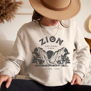 K&ouml;nnte beinhalten: Beiges Sweatshirt mit einem Grafikdesign des Zion National Park. Das Design enth&auml;lt den Text "ZION NATIONAL PARK" &uuml;ber einer Berg- und Baumillustration, mit "EST. 1919 UTAH" darunter. Das Sweatshirt hat eine neutrale Farbe.