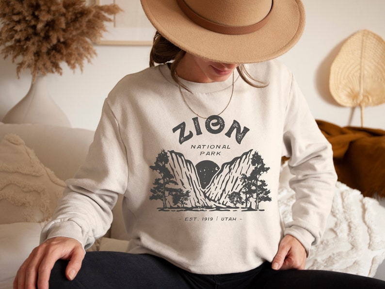 K&ouml;nnte beinhalten: Beiges Sweatshirt mit einem Grafikdesign des Zion National Park. Das Design enth&auml;lt den Text "ZION NATIONAL PARK" &uuml;ber einer Berg- und Baumillustration, mit "EST. 1919 UTAH" darunter. Das Sweatshirt hat eine neutrale Farbe.