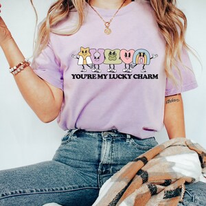 May include: A light purple t-shirt with a graphic of cartoon lucky charms spelling out "LUCKY" and the text "YOU'RE MY LUCKY CHARM".