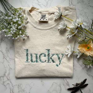 May include: A cream colored t-shirt with the word "lucky" embroidered in green leaves. The shirt is folded and laid on a white marble surface.