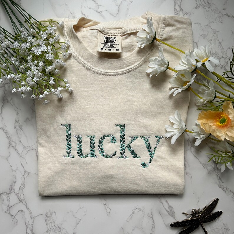 May include: A cream colored t-shirt with the word "lucky" embroidered in green leaves. The shirt is folded and laid on a white marble surface.