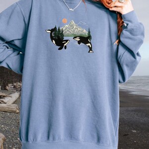 Puede incluir: Una sudadera azul claro con un gr&aacute;fico de dos orcas nadando frente a una cordillera con una puesta de sol en el fondo.