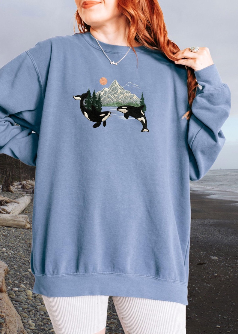 Puede incluir: Una sudadera azul claro con un gr&aacute;fico de dos orcas nadando frente a una cordillera con una puesta de sol en el fondo.