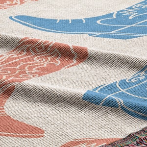 May include: A woven throw blanket with a cream background and a repeating pattern of blue and red cowboy boots. The blanket has a fringe border.