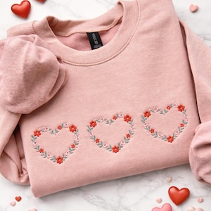 May include: A light pink sweatshirt featuring three embroidered heart-shaped floral designs. The hearts are outlined with green leaves and filled with red and pink flowers. Scattered around the sweatshirt are heart-shaped candies in red and pink.