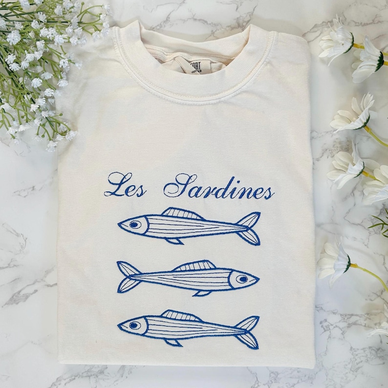 May include: A white t-shirt with a blue graphic of three sardines and the text "Les Sardines".