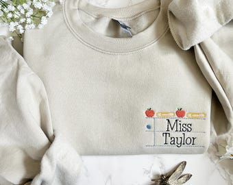 May include: A beige sweatshirt with a small embroidered design of a teacher's desk with apples and pencils. The text "Miss Taylor" is embroidered below the desk.