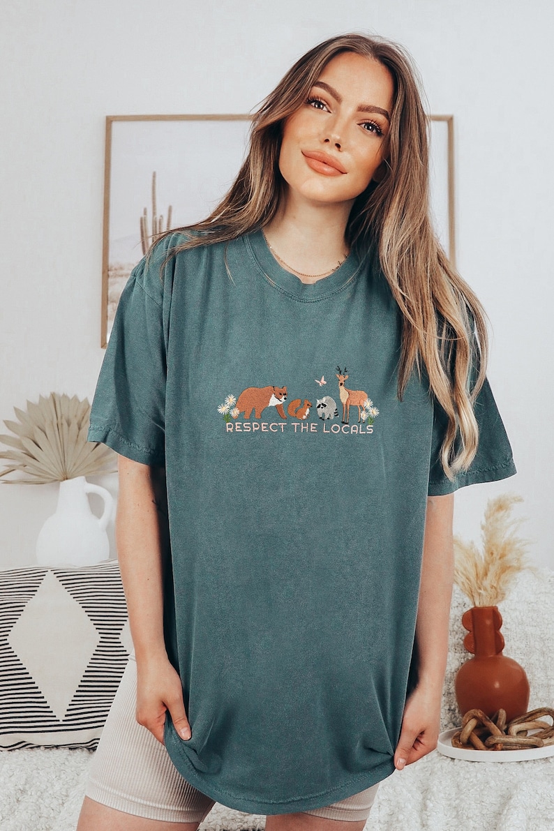 Puede incluir: Una camiseta azul verdosa con un gr&aacute;fico de un oso, un mapache, un ciervo y una mariposa con el texto "Respect the Locals".