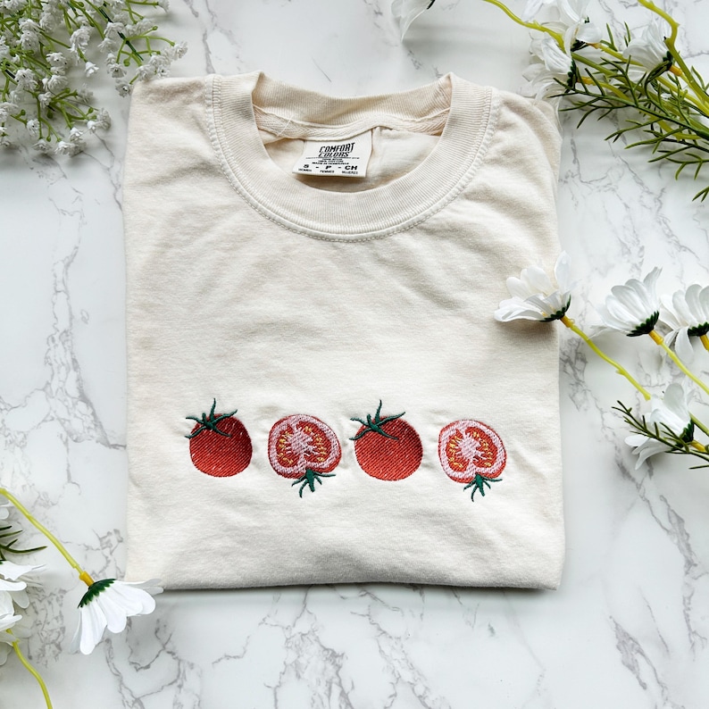 May include: Cream-colored t-shirt with embroidered red tomatoes. The design features three whole tomatoes and two sliced tomatoes with green stems. The shirt is folded and displayed on a marble surface.