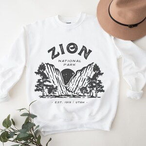 K&ouml;nnte beinhalten: Wei&szlig;es Sweatshirt mit schwarzer Grafik des Zion Nationalparks. Das Design enth&auml;lt den Text "ZION NATIONAL PARK" &uuml;ber einer Berg- und Baumillustration, mit "EST. 1919 | UTAH" darunter. Ein beiger Hut mit schwarzem Band liegt obenauf.