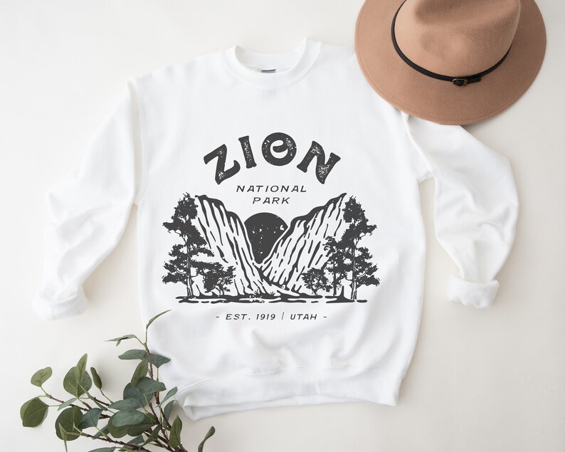 K&ouml;nnte beinhalten: Wei&szlig;es Sweatshirt mit schwarzer Grafik des Zion Nationalparks. Das Design enth&auml;lt den Text "ZION NATIONAL PARK" &uuml;ber einer Berg- und Baumillustration, mit "EST. 1919 | UTAH" darunter. Ein beiger Hut mit schwarzem Band liegt obenauf.