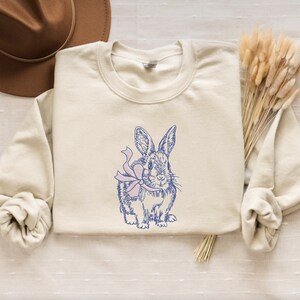 May include: A beige sweatshirt with a blue and pink illustration of a rabbit wearing a bow.