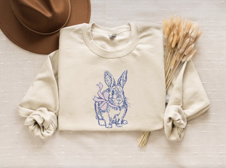 May include: A beige sweatshirt with a blue and pink illustration of a rabbit wearing a bow.