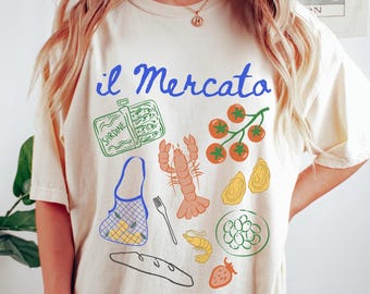 May include: Cream-colored t-shirt with the Italian phrase "il Mercato" in blue script. The shirt features colorful illustrations of sardines, a lobster, tomatoes, oysters, and other food items, evoking a market theme.