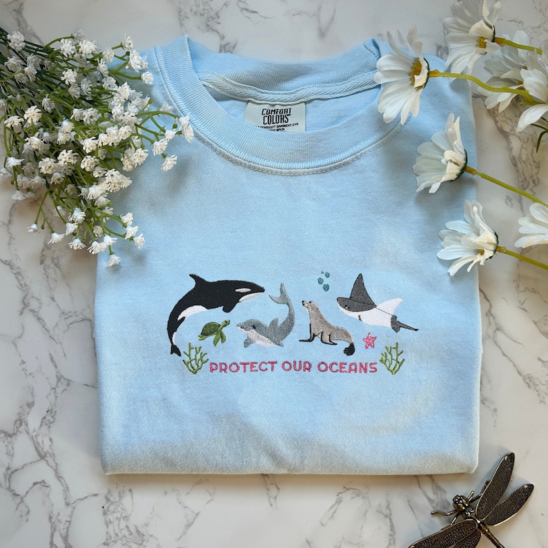 May include: Light blue t-shirt with an embroidered design of various sea creatures, including a whale, a turtle, a seal, and a stingray. The text "PROTECT OUR OCEANS" is embroidered below the sea creatures.